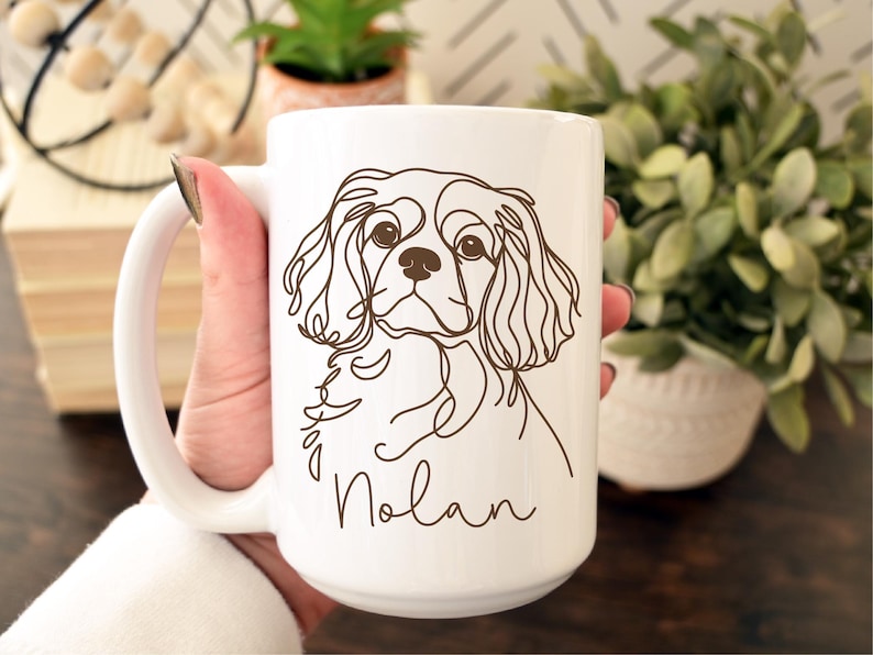 May include: White ceramic mug featuring a brown line drawing of a Cavalier King Charles Spaniel dog. The dog's name, "Nolan," is written in a script font below the dog's image. The mug has a large handle.