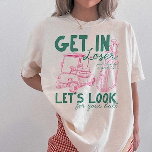 May include: Cream-colored t-shirt with a golf-themed graphic. The design features a golf cart and golf bag in pink, with the phrases "GET IN LOSER," "LET'S LOOK for your ball," and "just here for a good time" in green and pink.