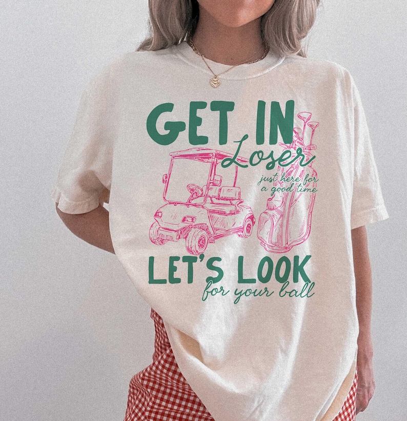 May include: Cream-colored t-shirt with a golf-themed graphic. The design features a golf cart and golf bag in pink, with the phrases "GET IN LOSER," "LET'S LOOK for your ball," and "just here for a good time" in green and pink.
