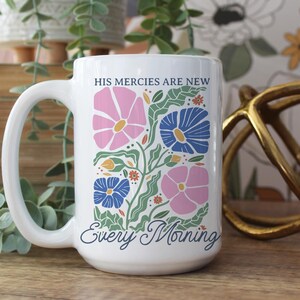 K&ouml;nnte beinhalten: Wei&szlig;er Keramikbecher mit gro&szlig;em Henkel, mit einem Blumendesign in Rosa, Blau und Gr&uuml;n. Der Becher tr&auml;gt den Text "HIS mercies are new" und "Every Morning". Der Becher steht auf einer Holzoberfl&auml;che.