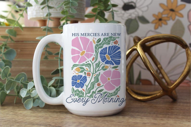 K&ouml;nnte beinhalten: Wei&szlig;er Keramikbecher mit gro&szlig;em Henkel, mit einem Blumendesign in Rosa, Blau und Gr&uuml;n. Der Becher tr&auml;gt den Text "HIS mercies are new" und "Every Morning". Der Becher steht auf einer Holzoberfl&auml;che.