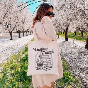 Puede incluir: Una bolsa de lona blanca con un dise&ntilde;o gr&aacute;fico negro. El dise&ntilde;o presenta una pila de libros, una botella de poci&oacute;n y el texto "Custom Name Book Club est. 2024".