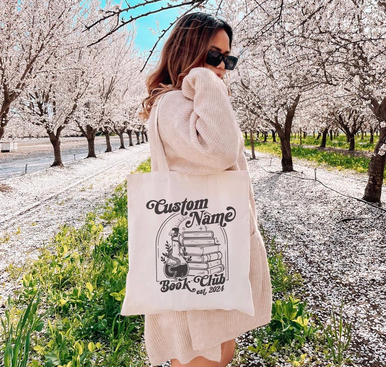 Puede incluir: Una bolsa de lona blanca con un dise&ntilde;o gr&aacute;fico negro. El dise&ntilde;o presenta una pila de libros, una botella de poci&oacute;n y el texto "Custom Name Book Club est. 2024".