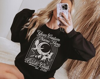 May include: Black sweatshirt with a white graphic design featuring a moon, mountains, and a dragon. The text reads "Your Custom Name Here" and "Book Club est. 2025". The sweatshirt has long sleeves.
