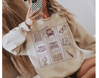 Puede incluir: Sudadera beige con una cuadrícula de nueve ilustraciones estilo sello postal. Las ilustraciones incluyen una pila de libros, un sillón, gafas, una taza de té, una máquina de escribir, un libro, un globo terráqueo, un libro abierto y una pluma. El diseño es de color marrón oscuro.