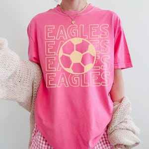 K&ouml;nnte beinhalten: Ein leuchtend pinkfarbenes T-Shirt mit kurzen &Auml;rmeln und einem gro&szlig;en, hellgelben Grafikdruck. Das Design besteht aus dem viermal untereinander gedruckten Wort EAGLES, wobei eine Fu&szlig;ballillustration zentral &uuml;ber dem Text platziert ist.