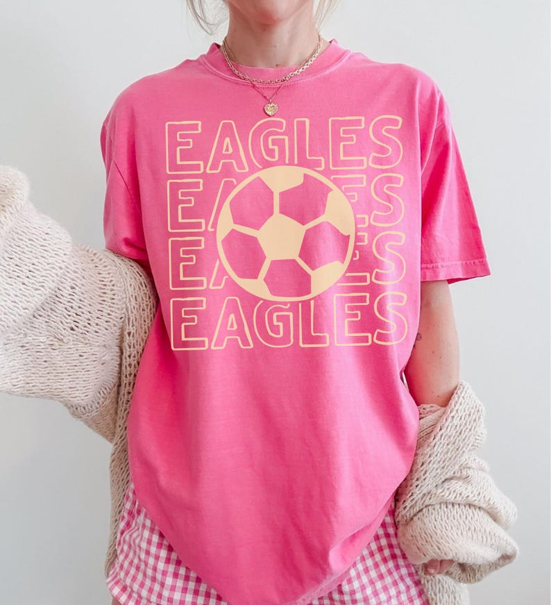 K&ouml;nnte beinhalten: Ein leuchtend pinkfarbenes T-Shirt mit kurzen &Auml;rmeln und einem gro&szlig;en, hellgelben Grafikdruck. Das Design besteht aus dem viermal untereinander gedruckten Wort EAGLES, wobei eine Fu&szlig;ballillustration zentral &uuml;ber dem Text platziert ist.