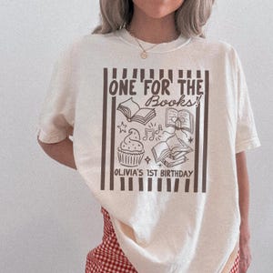Peut inclure: T-shirt crème avec un motif marron. Le motif comprend le texte "ONE FOR THE BOOKS !" ainsi que des illustrations de livres, un cupcake, des notes de musique et des étoiles. Le texte "OLIVIA'S 1ST BIRTHDAY" est également inclus.