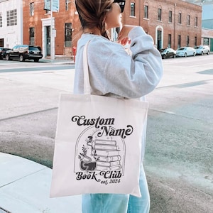 Puede incluir: Una bolsa de lona blanca con texto negro y un dise&ntilde;o gr&aacute;fico. El texto dice "Custom Name Book Club est. 2024". El dise&ntilde;o gr&aacute;fico presenta una pila de libros con una botella de poci&oacute;n y una serpiente.