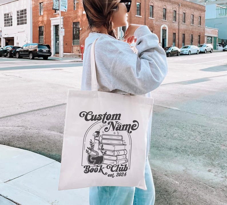 Puede incluir: Una bolsa de lona blanca con texto negro y un dise&ntilde;o gr&aacute;fico. El texto dice "Custom Name Book Club est. 2024". El dise&ntilde;o gr&aacute;fico presenta una pila de libros con una botella de poci&oacute;n y una serpiente.