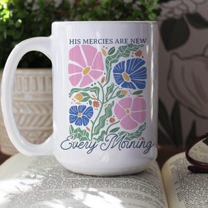 K&ouml;nnte beinhalten: Wei&szlig;e Keramiktasse mit einem Blumendesign in Rosa, Blau und Gr&uuml;n. Die Tasse tr&auml;gt den Text "HIS MERCIES ARE NEW" und "Every Morning". Die Tasse steht auf einem offenen Buch.