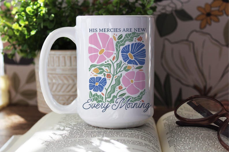 K&ouml;nnte beinhalten: Wei&szlig;e Keramiktasse mit einem Blumendesign in Rosa, Blau und Gr&uuml;n. Die Tasse tr&auml;gt den Text "HIS MERCIES ARE NEW" und "Every Morning". Die Tasse steht auf einem offenen Buch.