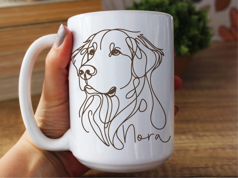 May include: White ceramic mug featuring a brown line drawing of a dog's face. The dog's name, "Nora", is written in a cursive font below the drawing. The mug has a large handle and is held by a person.