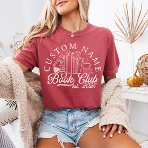 May include: A dusty rose t-shirt with a white graphic that reads "CUSTOM NAME Book Club est. 2025." The design includes books, flowers, and a coffee cup. The shirt is paired with denim shorts.