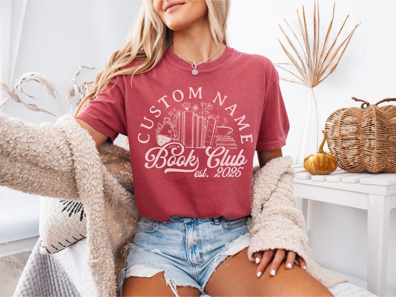 May include: A dusty rose t-shirt with a white graphic that reads "CUSTOM NAME Book Club est. 2025." The design includes books, flowers, and a coffee cup. The shirt is paired with denim shorts.