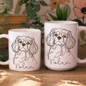 May include: Two white ceramic mugs with a line drawing of a Cavalier King Charles Spaniel. The dog's name, "Nolan", is written in cursive below the drawing. The mugs are on a wooden surface.