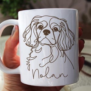 May include: White ceramic mug featuring a brown line drawing of a Cavalier King Charles Spaniel dog. The dog's name, "Nolan", is written in a cursive font below the dog's image. The mug has a white handle.