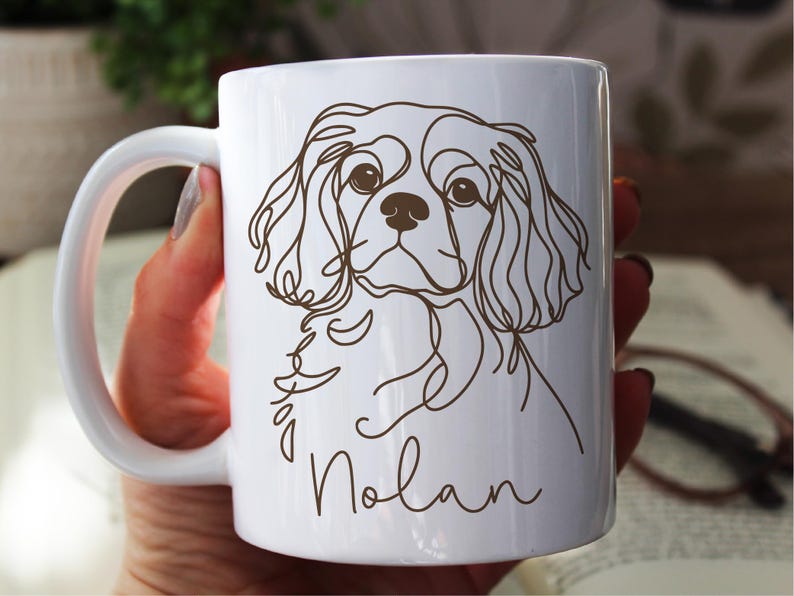 May include: White ceramic mug featuring a brown line drawing of a Cavalier King Charles Spaniel dog. The dog's name, "Nolan", is written in a cursive font below the dog's image. The mug has a white handle.