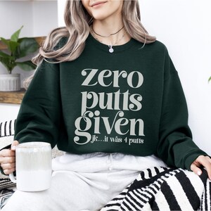 May include: A dark green sweatshirt with the text "zero putts given" in white, and the text "jk... it was 4 putts" in smaller white text below.