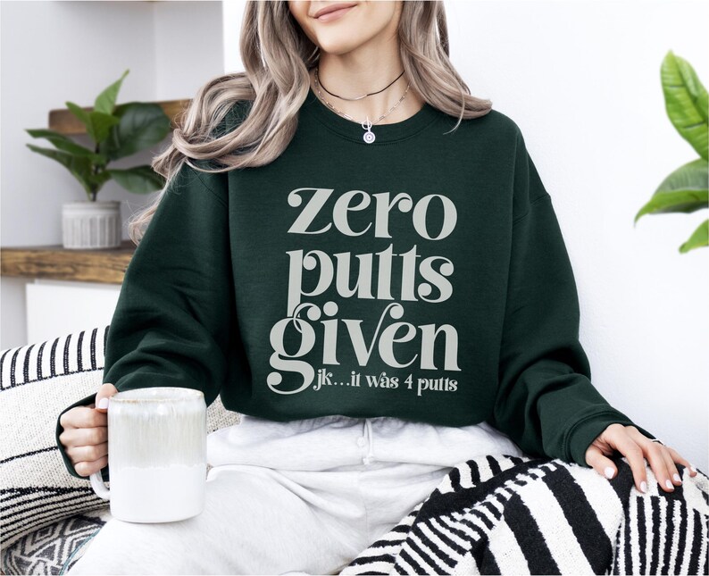 May include: A dark green sweatshirt with the text "zero putts given" in white, and the text "jk... it was 4 putts" in smaller white text below.