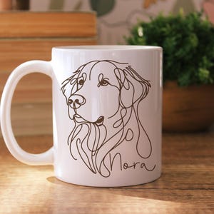 May include: White ceramic mug featuring a line drawing of a dog's face in brown. The dog's name, "Nora", is written in cursive below the drawing. The mug has a curved handle and is set against a wooden surface.