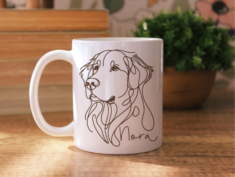May include: White ceramic mug featuring a line drawing of a dog's face in brown. The dog's name, "Nora", is written in cursive below the drawing. The mug has a curved handle and is set against a wooden surface.