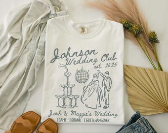 May include: White t-shirt with a gray graphic design that says "Johnson Wedding Club est. 2025" with a drawing of a couple dancing and a champagne tower. The text below the graphic says "Josh & Maggie's Wedding I CAME - I DRANK - I GOT A HANGOVER"