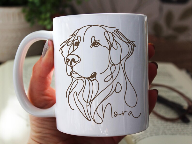 May include: White ceramic mug featuring a brown line drawing of a dog's head and the name "Nora" in a script font. The mug has a curved handle and is held by a person. The design is minimalist and elegant.