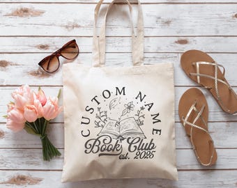 Puede incluir: Un bolso tote color crema con la inscripción "CUSTOM NAME Book Club est. 2025" en negro. La imagen incluye gafas de sol, sandalias y flores rosas, dispuestas sobre una superficie de madera clara.