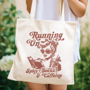 May include: A white canvas tote bag with a red graphic design. The graphic features a woman with red sunglasses reading a book and holding a glass of iced coffee. The text on the bag reads "Running on Spicy Books & Caffeine".