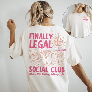 May include: Cream-coloured t-shirt with a pink graphic design on the back that reads "FINALLY LEGAL SOCIAL CLUB" with a martini glass, bow, and disco ball. The front of the shirt has a smaller graphic that says "Birthday Girl".