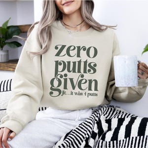 May include: A beige sweatshirt with the text "Zero putts given...jk...it was 4 putts" in dark green letters.