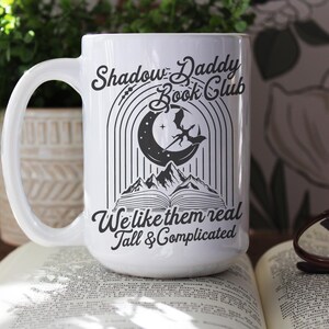 May include: White ceramic mug with a black graphic design. The design features a crescent moon, a dragon, and a book. The text reads "Shadow Daddy Book Club We like them real Tall & Complicated".