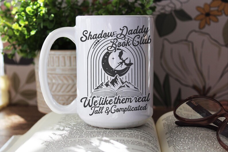 May include: White ceramic mug with a black graphic design. The design features a crescent moon, a dragon, and a book. The text reads "Shadow Daddy Book Club We like them real Tall & Complicated".