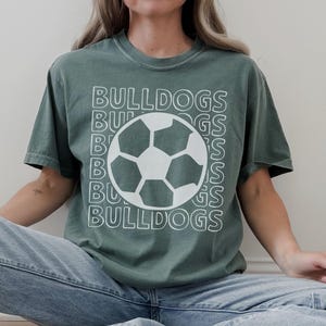 K&ouml;nnte beinhalten: Ein salbeigr&uuml;nes Kurzarm-T-Shirt mit einem wei&szlig;en Grafikdruck eines Fu&szlig;balls, der &uuml;ber dem Wort BULLDOGS zentriert ist, welches in einem gestapelten, umrandeten Schriftmuster wiederholt wird. Das Shirt hat einen Rundhalsausschnitt und eine lockere Passform.