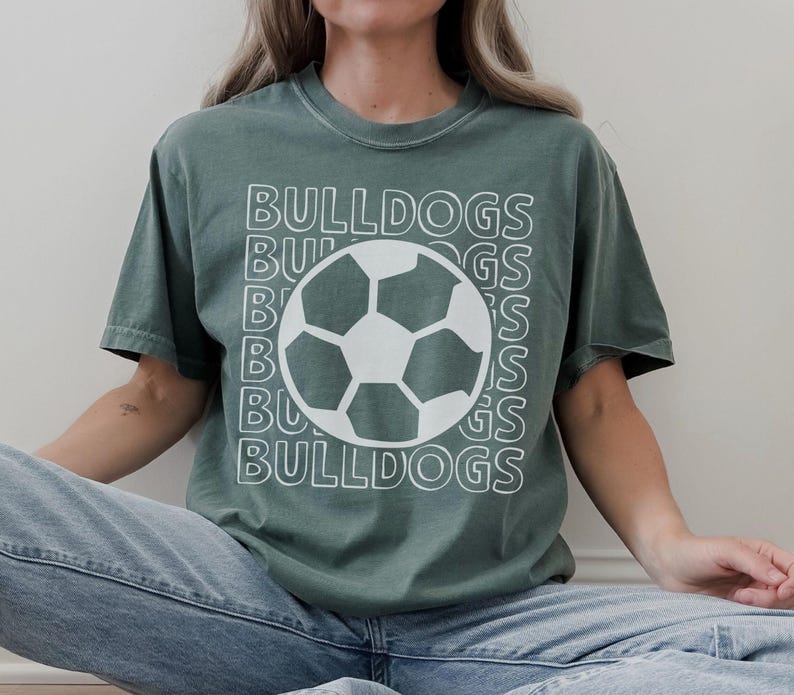 K&ouml;nnte beinhalten: Ein salbeigr&uuml;nes Kurzarm-T-Shirt mit einem wei&szlig;en Grafikdruck eines Fu&szlig;balls, der &uuml;ber dem Wort BULLDOGS zentriert ist, welches in einem gestapelten, umrandeten Schriftmuster wiederholt wird. Das Shirt hat einen Rundhalsausschnitt und eine lockere Passform.