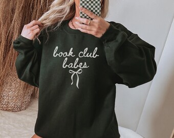 May include: Dark green sweatshirt with the text "book club babes" in white script. A white bow graphic is below the text. The sweatshirt has long sleeves and a crew neck. The image shows a person wearing the sweatshirt.