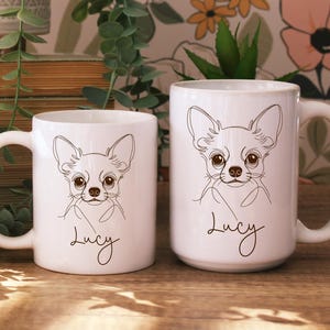 May include: Two white ceramic mugs with a line drawing of a Chihuahua dog. The dog has large eyes and ears, and the name "Lucy" is written below the drawing in a cursive font. The mugs are on a wooden surface.