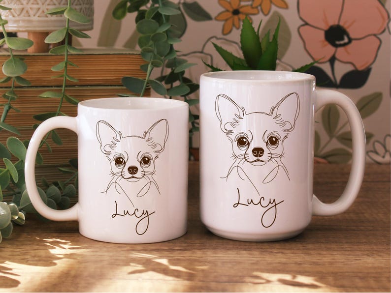 May include: Two white ceramic mugs with a line drawing of a Chihuahua dog. The dog has large eyes and ears, and the name "Lucy" is written below the drawing in a cursive font. The mugs are on a wooden surface.
