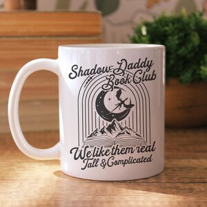 May include: White ceramic mug with a black design featuring a crescent moon, a dragon, and a book. The text reads "Shadow Daddy Book Club We like them real Tall & Complicated".