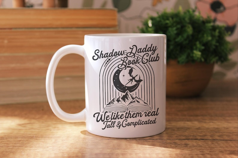 May include: White ceramic mug with a black design featuring a crescent moon, a dragon, and a book. The text reads "Shadow Daddy Book Club We like them real Tall & Complicated".