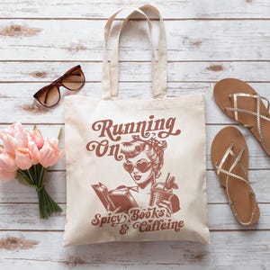 May include: A white tote bag with a brown design featuring a woman with sunglasses and the text "Running on Spicy Books & Caffeine".