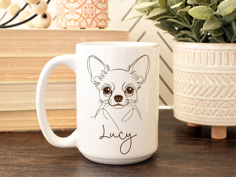 May include: White ceramic mug featuring a line drawing of a Chihuahua's face. The dog's name, "Lucy," is written in cursive below the image. The mug has a large handle and is suitable for hot or cold beverages.