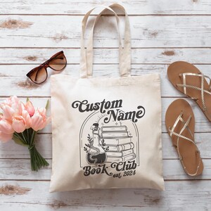 Puede incluir: Una bolsa de tela blanca con texto negro y un gr&aacute;fico de una botella de poci&oacute;n y libros apilados. El texto dice "Custom Name Book Club est. 2024".