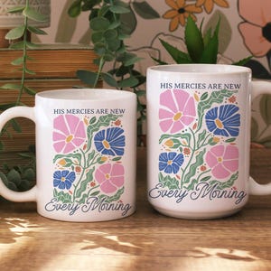 K&ouml;nnte beinhalten: Zwei wei&szlig;e Keramikbecher mit floralen Designs. Die Becher zeigen rosa und blaue Blumen mit gr&uuml;nen Bl&auml;ttern und den Text "HIS MERCIES ARE NEW Every Morning". Die Becher stehen auf einer Holzoberfl&auml;che.