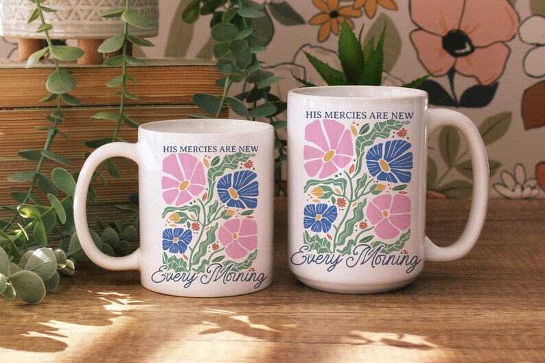K&ouml;nnte beinhalten: Zwei wei&szlig;e Keramikbecher mit floralen Designs. Die Becher zeigen rosa und blaue Blumen mit gr&uuml;nen Bl&auml;ttern und den Text "HIS MERCIES ARE NEW Every Morning". Die Becher stehen auf einer Holzoberfl&auml;che.