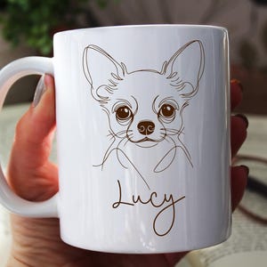 May include: White ceramic mug featuring a line drawing of a Chihuahua's face in brown. The dog's name, "Lucy", is written in a cursive font below the drawing. The mug has a white handle and is held by a person.