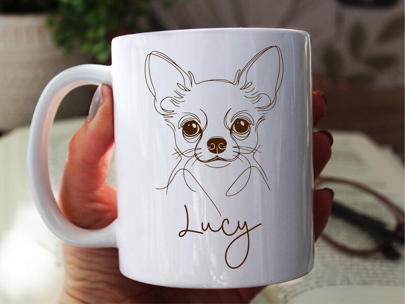 May include: White ceramic mug featuring a line drawing of a Chihuahua's face in brown. The dog's name, "Lucy", is written in a cursive font below the drawing. The mug has a white handle and is held by a person.