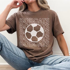 K&ouml;nnte beinhalten: Ein braunes Kurzarm-T-Shirt mit dem Wort BOBCATS, das in wei&szlig;er Umriss-Schrift auf der Vorderseite wiederholt wird, zentriert mit einer gro&szlig;en wei&szlig;en und braunen Fu&szlig;ballgrafik in der Mitte des Textdesigns.