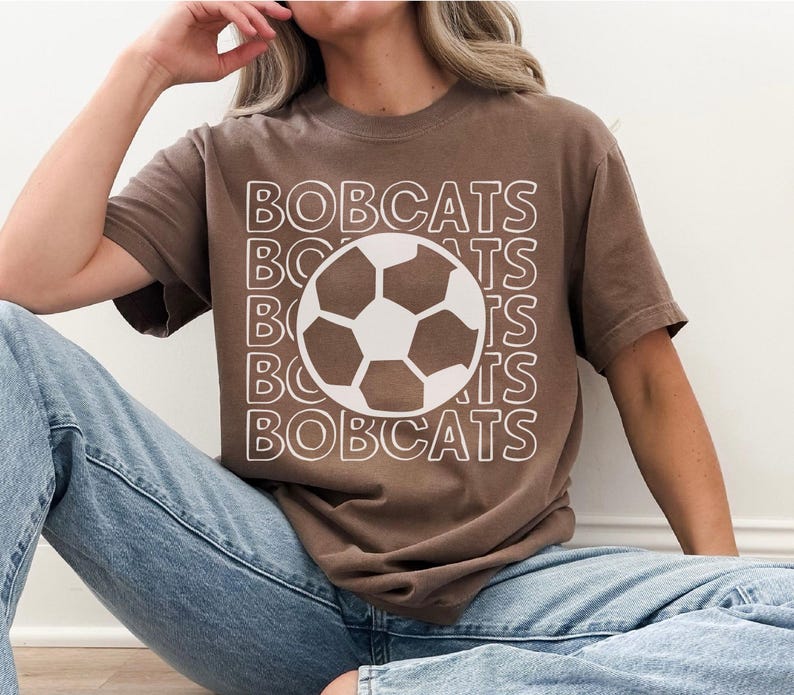K&ouml;nnte beinhalten: Ein braunes Kurzarm-T-Shirt mit dem Wort BOBCATS, das in wei&szlig;er Umriss-Schrift auf der Vorderseite wiederholt wird, zentriert mit einer gro&szlig;en wei&szlig;en und braunen Fu&szlig;ballgrafik in der Mitte des Textdesigns.